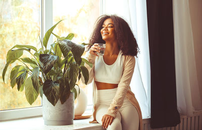 Smiling teenager girl drinking water sitting by window