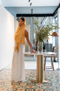 Full body of charming muslim female in traditional hijab looking at camera while standing near table with flowers in room