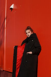 Fashionable teenage girl wearing black warm clothing standing against red wall during sunny day