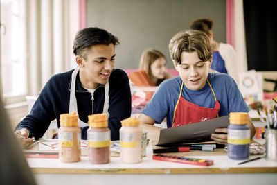 Smiling teenage boy showing artwork to male friend during painting class at high school