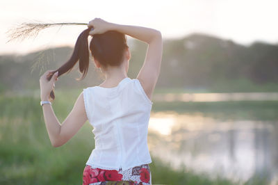 Rear view of woman tying up hair while standing on field during sunset