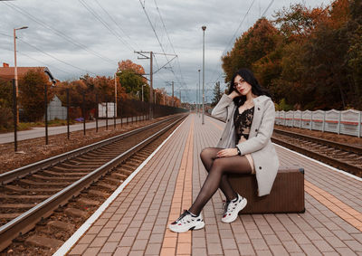 Young woman sitting on railroad track against sky