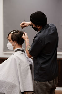 Barber in protective mask and latex glove styling adult man in a barbershop salon. new normal