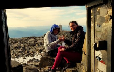 Mother and daughter bumping fists while sitting on rock against sky