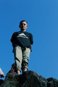 Low angle view of man standing on rock against clear blue sky