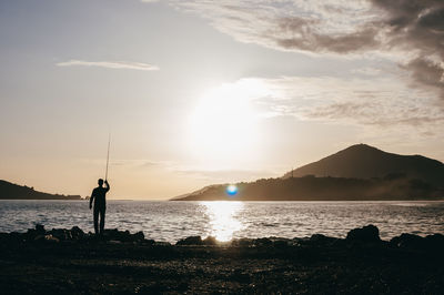 Silhouette person fishing in sea against sky during sunset