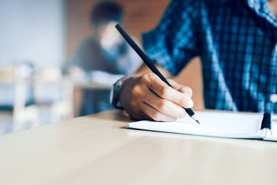 Midsection of man writing in book on table at classroom