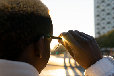 Side view of crop unrecognizable african american male putting on stylish sunglasses while standing on sunny street in city with buildings