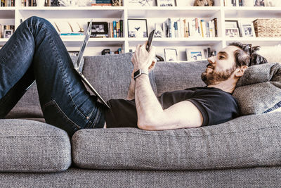 Man working with laptop computer and smart phone while laying on home sofa couch.
