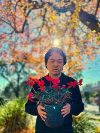 Portrait of young asian man holding red flowering cyclamen potted plant against autumn foliage 