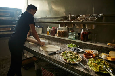 Side view of man using rolling pin to stretch fresh dough on on marble counter table while preparing pizza in kitchen