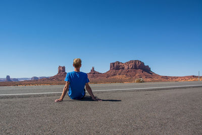 Rear view of man sitting on desert against clear blue sky