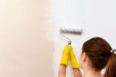 Teenage girl paints a wall with a roller of white paint, in yellow gloves