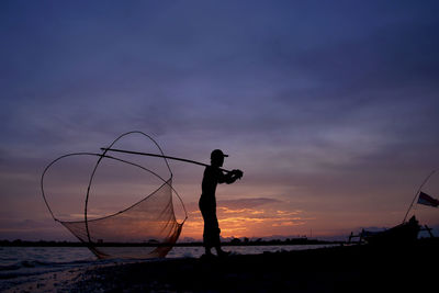 Silhouette fisherman on beach against sky during sunset