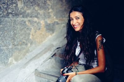 Portrait of smiling woman holding camera while sitting on staircase