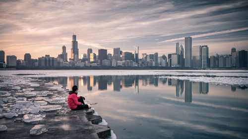 Man sitting at riverbank during winter in city