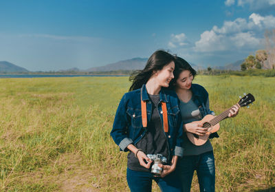 Young women standing on grass while holding ukulele and camera against sky
