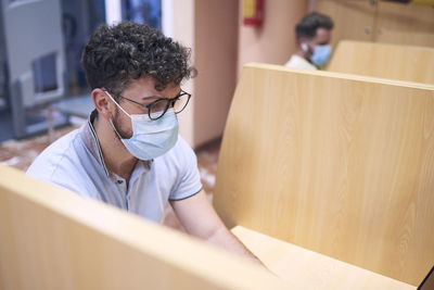 A boy wearing a mask is studying in a personal cabin in the library