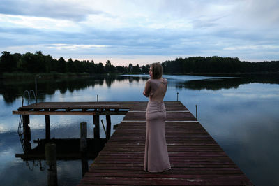 Rear view of young woman wearing gown standing on pier over lake against sky during sunset