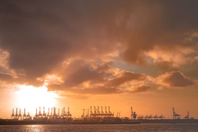 Cranes at harbor against cloudy sky during sunset