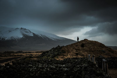 Scenic view of person standing on hill against snow covered mountains and dramatic sky 