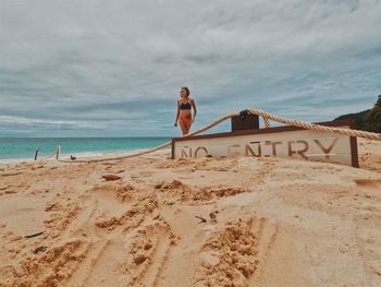 Woman in bikini walking on shore by no entry text at beach against cloudy sky