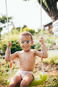 Portrait of shirtless boy sitting on swing against sky
