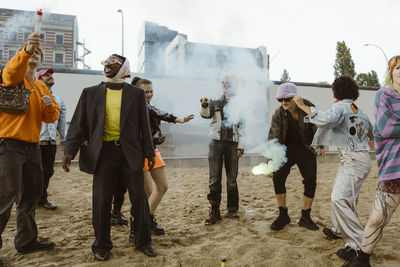 Carefree non-binary friends burning distress flare while standing on sand at park