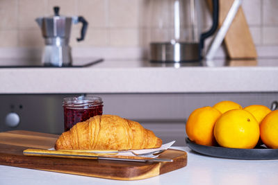 Wooden serving board with croissant and jam and dish of oranges lies on kitchen table in kitchen