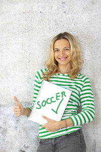 Portrait of smiling beautiful woman holding book with text while standing against wall