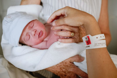 Happy mature female greeting newborn grandchild in hospital ward while young mother lying in bed and receiving medical treatment