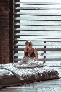 Portrait of young woman sitting on bed against window