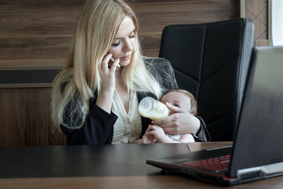 Woman feeds baby from bottle  and talks on phone, concept of combining business and caring for baby.