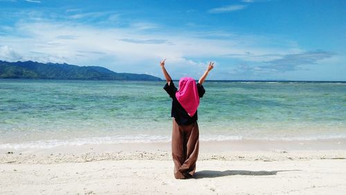 Rear view of woman with arms raised walking at beach against sky