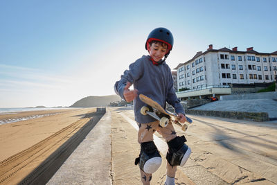 Cheerful teenage boy in helmet standing with skateboard on promenade near sea in summer