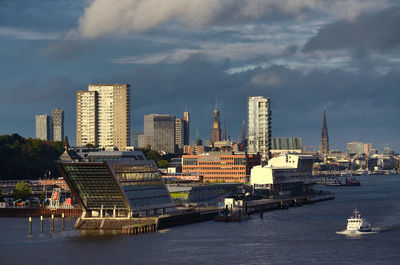 Modern buildings by river against cloudy sky in city