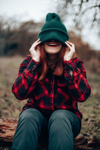 Happy young girl in a green knit cap pulled over her eyes and a plaid shirt in the autumn forest