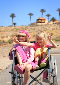 Cute siblings sitting on wheelchair against sky