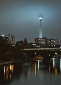 Illuminated bridge over river by buildings against sky at night