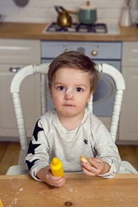 Boy sitting in the kitchen at the table with a rolling pin and flour on his nose