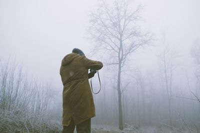 Rear view of man standing by bare tree in foggy weather