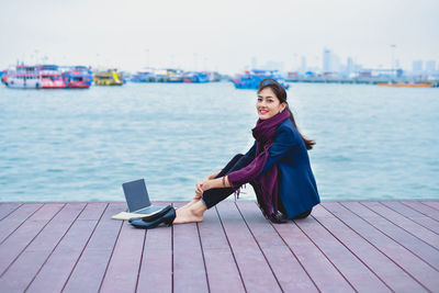 Full length portrait of smiling woman sitting on pier over sea