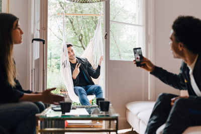 Man photographing friend with smart phone while sitting in swing at home