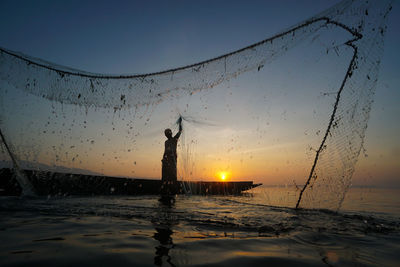 Silhouette fisherman standing by sea against sky during sunset