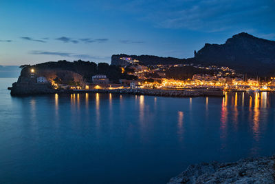 Illuminated buildings by sea against sky at dusk