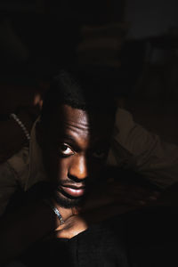 Confident young african american guy relaxing in dark room near window with sunlight on face and looking at camera