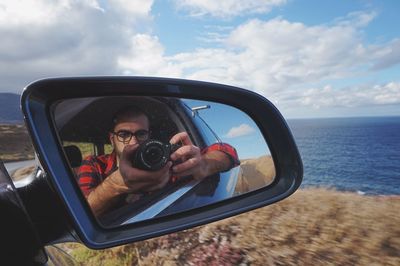 Man photographing self in side-view mirror against sky