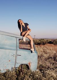 Full length of woman sitting on abandoned van at field against clear sky