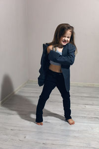 Funny portrait of fashionable girl child in large gray jacket stands against gray wall next  window 