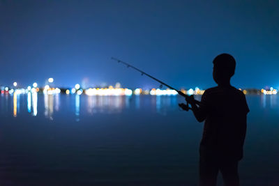 Rear view of silhouette boy fishing in sea at night